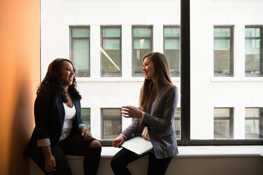 Two businesswomen enjoying a conversation by a large office window.