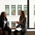 Two businesswomen enjoying a conversation by a large office window.