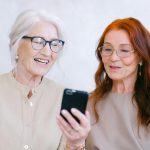 Two senior women sharing a moment of laughter while using a smartphone indoors.