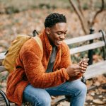 Side view of young positive African American man with backpack sitting in park on autumn day and using modern smartphone