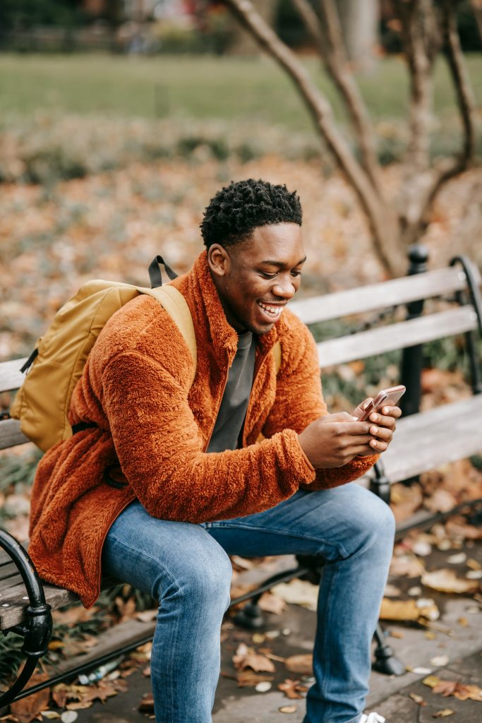 Side view of young positive African American man with backpack sitting in park on autumn day and using modern smartphone