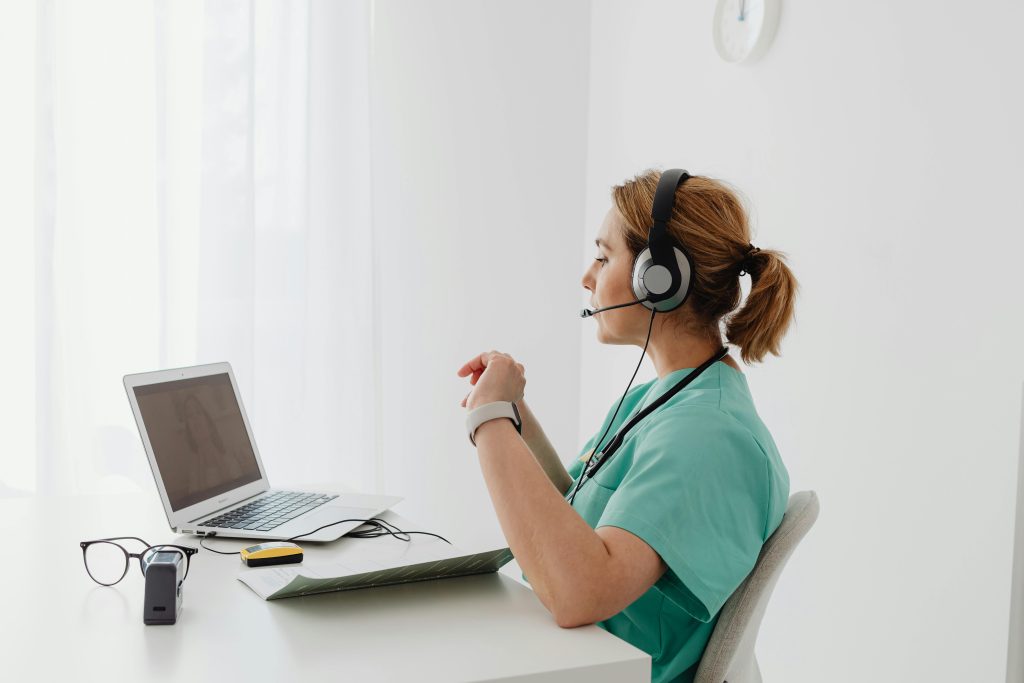 A female doctor using a laptop for an online consultation, wearing a headset in a bright office.