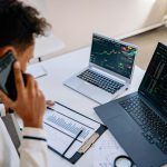 Man analyzing stock market charts on laptops while talking on a cellphone.