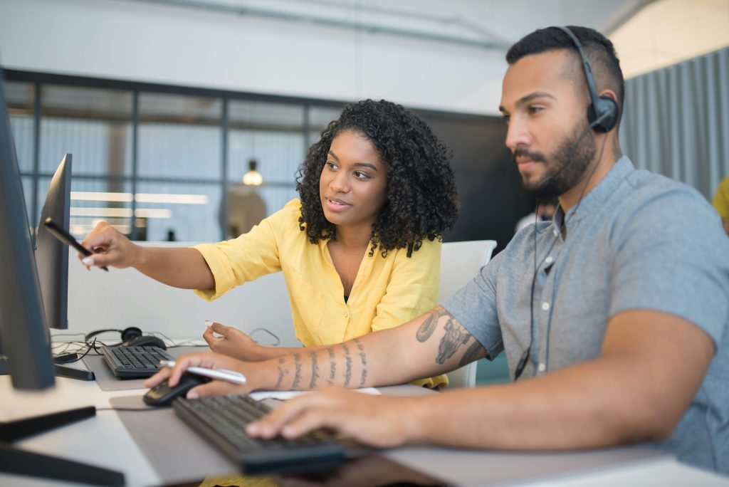 Two colleagues working together at a computer, discussing solutions with headsets in a modern office.