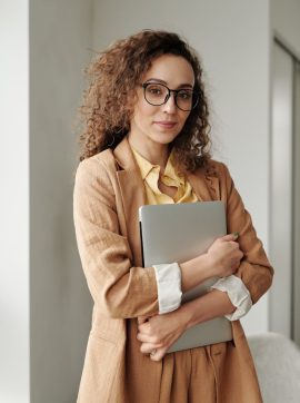 A confident woman in formal attire holding a laptop, standing in an office environment and looking at the camera.