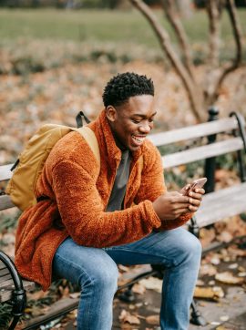 Side view of young positive African American man with backpack sitting in park on autumn day and using modern smartphone